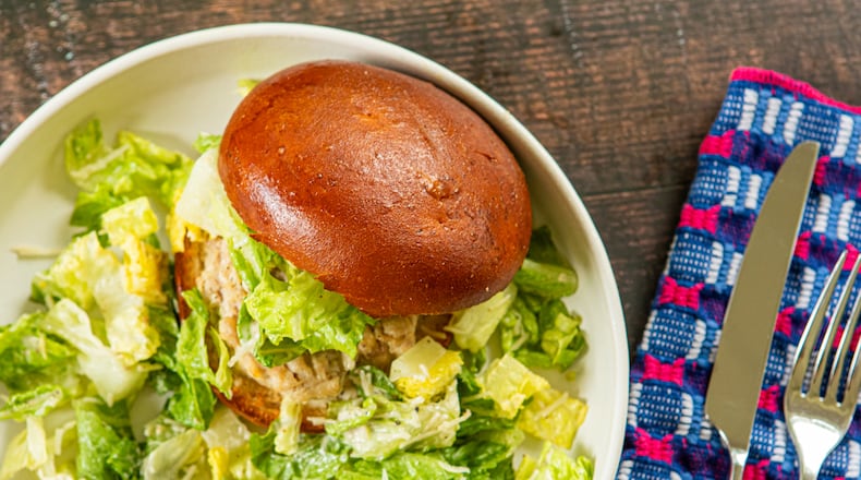 A plate filled with a chicken burger on a smooth bun garnished with Caesar salad, and also with Caesar salad on the side. The plate is on a wooden table top with a fork, knife and napkin next to it.