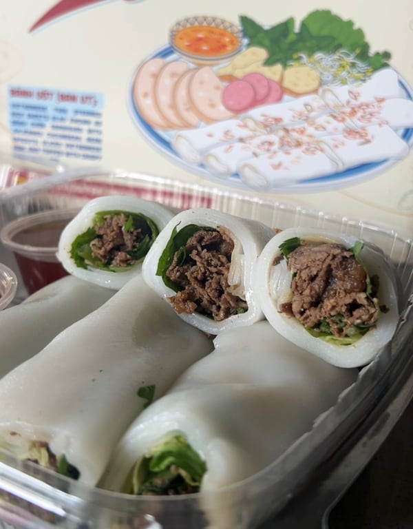 Six beef rolls, called pho bo cuon, in a plastic take-out container with a side of dipping sauce.