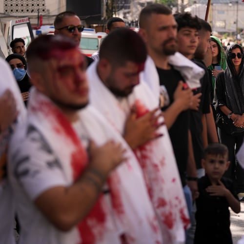 A woman watches as Lebanese Shiites beat their chests and bleed from self-inflicted ceremonial head wounds during Ashoura, when Shiites commemorate the martyrdom of Imam Hussein, the grandson of the Prophet Muhammad, amid buildings destroyed by Israeli strikes in Nabatiyeh, southern Lebanon, Friday, June 26, 2026. (AP Photo/Hassan Ammar)