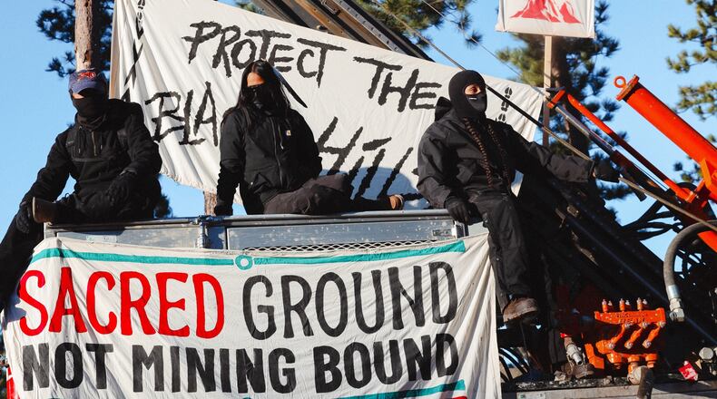 Masked demonstrators sit atop drilling equipment on Thursday, April 30, 2026, in Pennington County, S.D., with a banner reading Sacred ground not mining bound. (Angel White Eyes via AP)