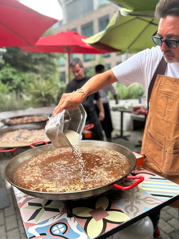 Man with apron and sunglasses pouring a steam pan filled with uncooked rice into a paella pan filled with broth.