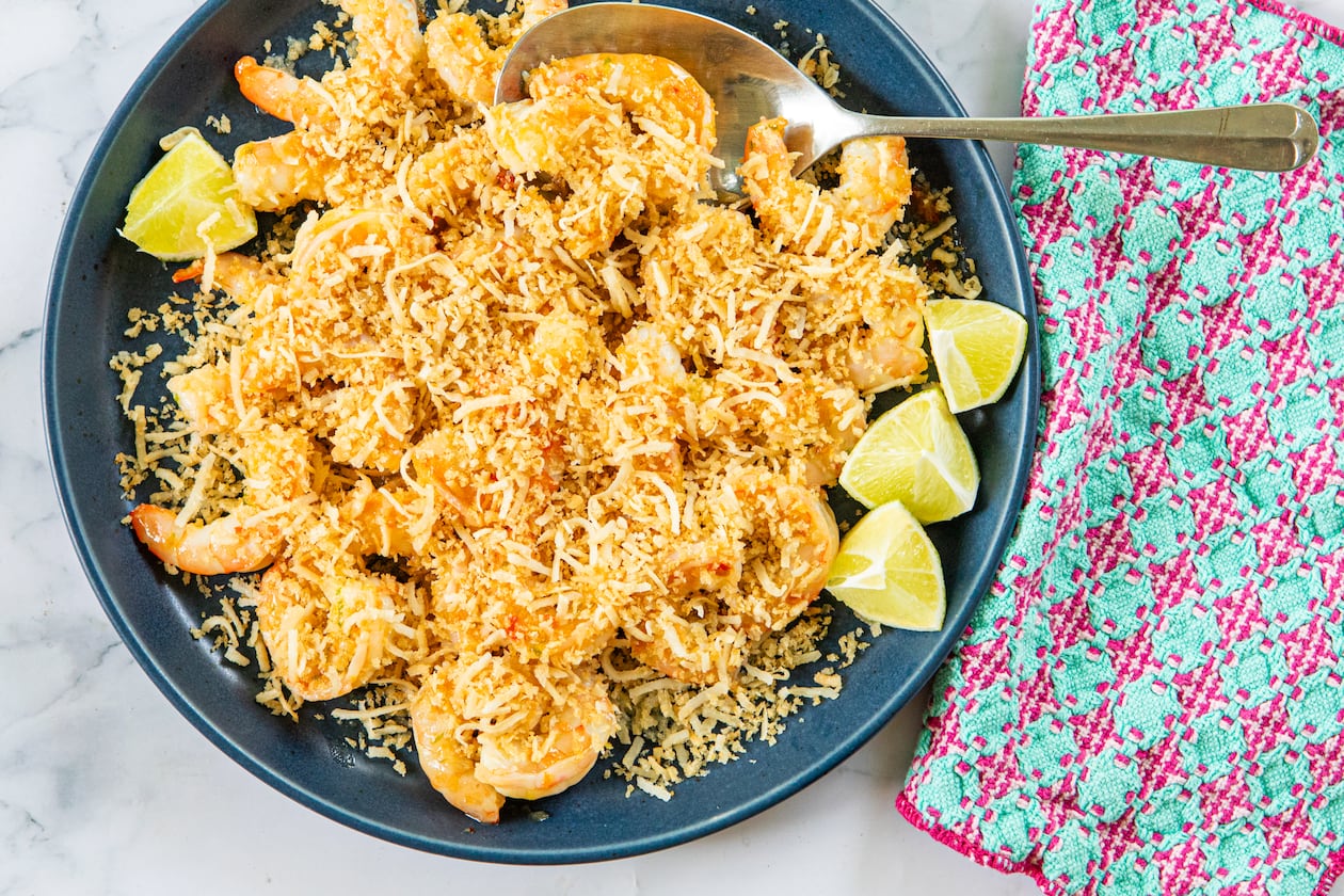 A serving dish is filled with sauteed shrimp covered by a topping made of panko and shaved coconut. A serving spoon is tucked into the bowl, and a colorful napkin is draped alongside the dish. All are arranged on a marble tabletop.