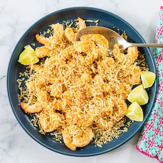 A serving dish is filled with sauteed shrimp covered by a topping made of panko and shaved coconut. A serving spoon is tucked into the bowl, and a colorful napkin is draped alongside the dish. All are arranged on a marble tabletop.