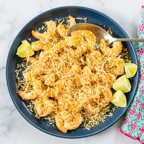 A serving dish is filled with sauteed shrimp covered by a topping made of panko and shaved coconut. A serving spoon is tucked into the bowl, and a colorful napkin is draped alongside the dish. All are arranged on a marble tabletop.