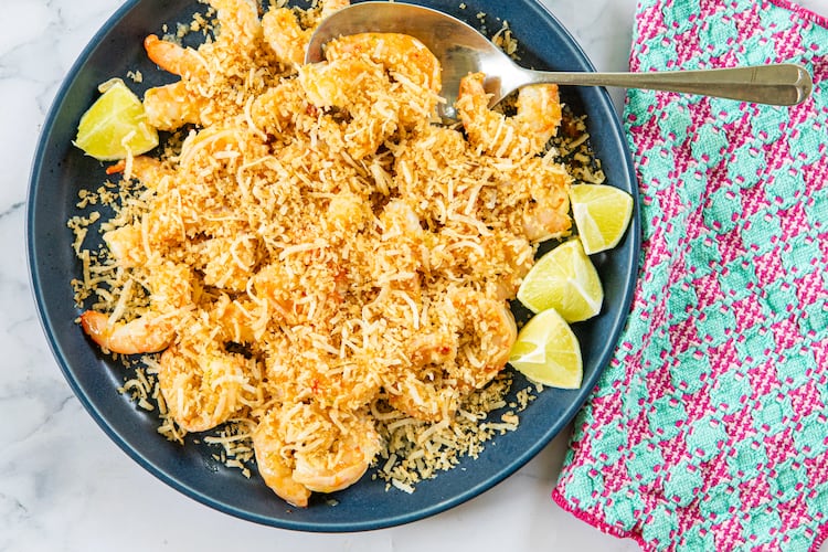 A serving dish is filled with sauteed shrimp covered by a topping made of panko and shaved coconut. A serving spoon is tucked into the bowl, and a colorful napkin is draped alongside the dish. All are arranged on a marble tabletop.