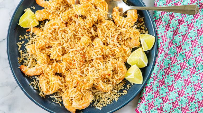 A serving dish is filled with sauteed shrimp covered by a topping made of panko and shaved coconut. A serving spoon is tucked into the bowl, and a colorful napkin is draped alongside the dish. All are arranged on a marble tabletop.