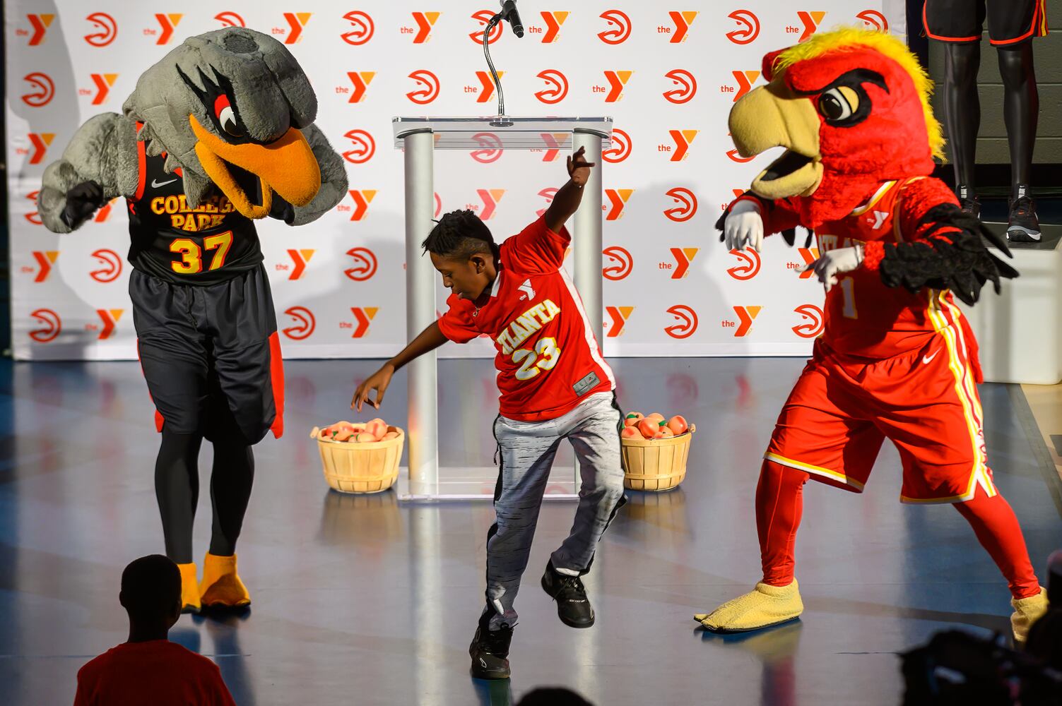 Collie Hawk, College Park Skyhawk's mascot, and Harry, the Atlanta Hawks mascot celebrate with one of the local YMCA students at the new sponsorship patch unveiling at The Villages at Carver Family YMCA in Atlanta, Georgia, November 20, 2023. (Jamie Spaar for the Atlanta Journal Constitution)