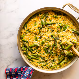 A large pot of creamy small pasta (orzo) and pieces of spring asparagus is sitting on a marble countertop. A wooden spoon is resting against the edge of the pot, and a towel is wrapped around the handle of the pot.