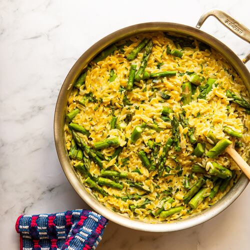 A large pot of creamy small pasta (orzo) and pieces of spring asparagus is sitting on a marble countertop. A wooden spoon is resting against the edge of the pot, and a towel is wrapped around the handle of the pot.