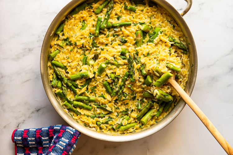 A large pot of creamy small pasta (orzo) and pieces of spring asparagus is sitting on a marble countertop. A wooden spoon is resting against the edge of the pot, and a towel is wrapped around the handle of the pot.
