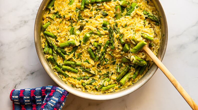 A large pot of creamy small pasta (orzo) and pieces of spring asparagus is sitting on a marble countertop. A wooden spoon is resting against the edge of the pot, and a towel is wrapped around the handle of the pot.