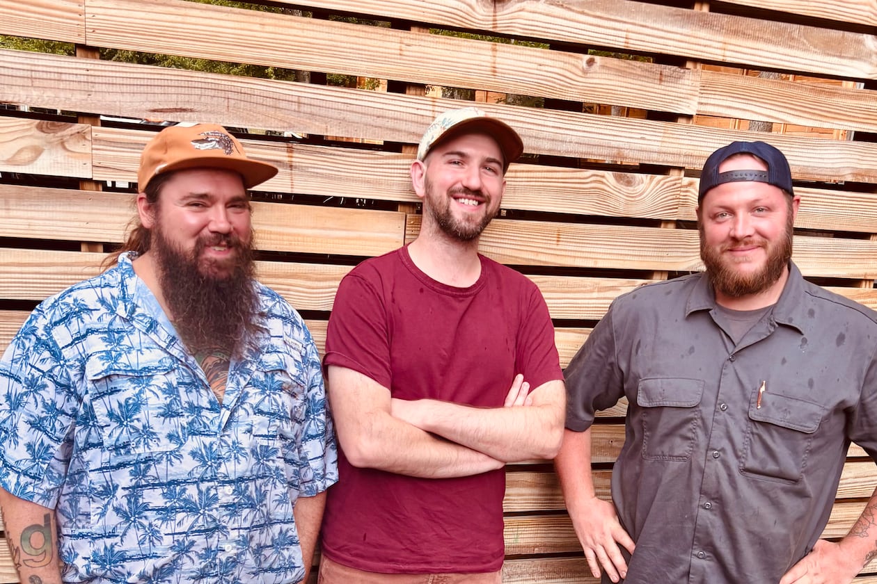 Three smiling men in nice casual clothing standing in front of a wooden-slat fence.