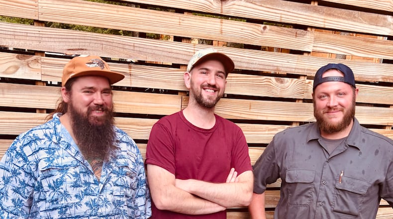 Three smiling men in nice casual clothing standing in front of a wooden-slat fence.
