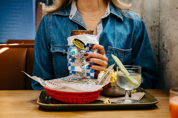 A woman in a denim jacket holds a foil-wrapped pastry pocket labeled with as lemon pepper flavor. A tray sits in front of her with a red fast food basket and a clear cocktail garnished with a long piece of celery.