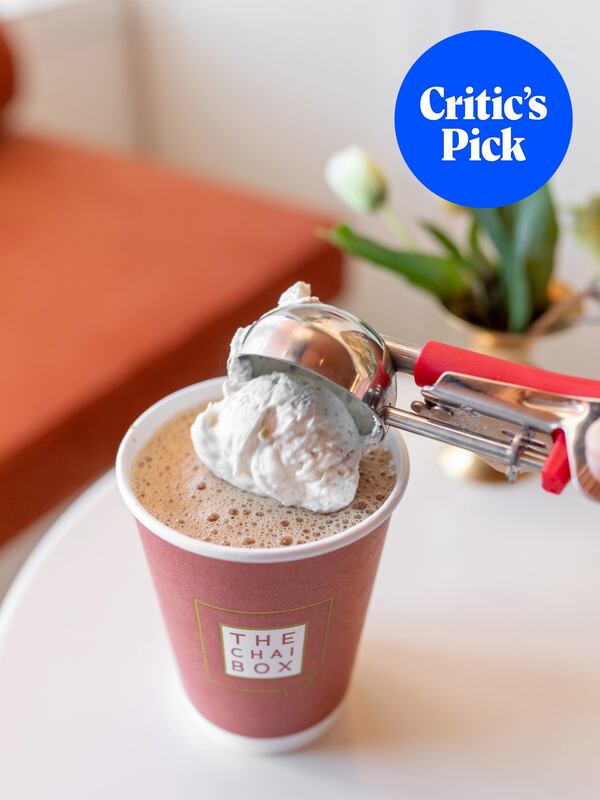 A paper cup labeled the Chai Box is full of hot chai with an ice cream scoop depositing a scoop of cream on top