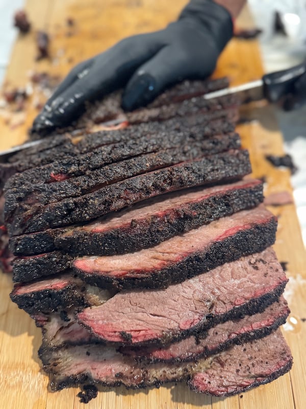 A gloved hand slices a smoked brisket with a blackened exterior into thick pieces.