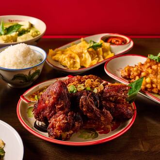 A group of Thai dishes on a restaurant table, with a heavily seasoned half-chicken cut into pieces on a plate in the center, surrounded by papaya salad, curry, a bowl of soup, a bowl of rice, a plate of dumplings and a fried appetizer.