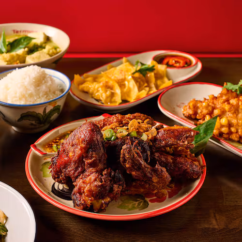 A group of Thai dishes on a restaurant table, with a heavily seasoned half-chicken cut into pieces on a plate in the center, surrounded by papaya salad, curry, a bowl of soup, a bowl of rice, a plate of dumplings and a fried appetizer.