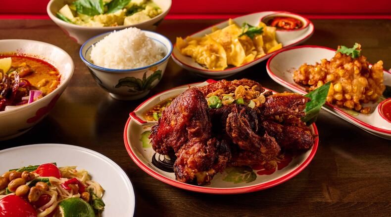 A group of Thai dishes on a restaurant table, with a heavily seasoned half-chicken cut into pieces on a plate in the center, surrounded by papaya salad, curry, a bowl of soup, a bowl of rice, a plate of dumplings and a fried appetizer.
