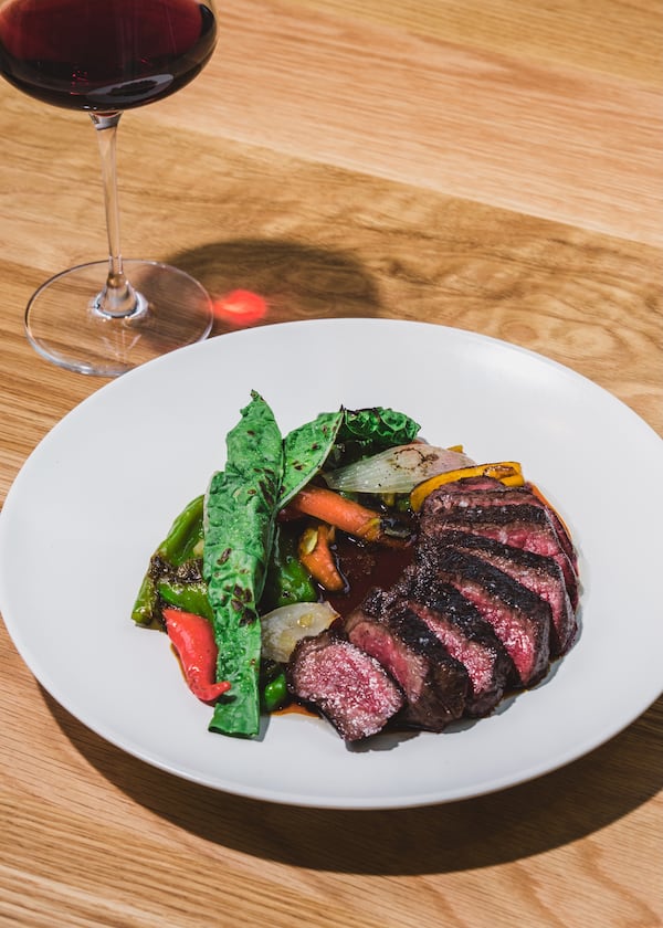 A plate of sliced steak arranged so that it curls around the vegetables accompanying it with a glass of wine on the left side.