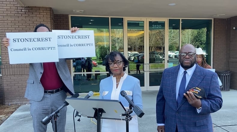 Michael Fayoyin, left, holds signs that read, "Stonecrest Council is Corrupt!!" during the news conference held by mayoral candidates Diane Adoma (center) and Kirby Frazier (right).