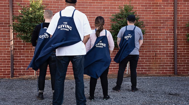 Four members of the Giving Kitchen staff are pictured facing away from the camera, wearing their aprons like capes.