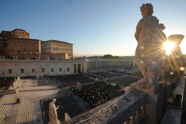 The sun rises through a statue as people begin to take their seats in St. Peter's Square, ahead of the funeral of Pope Francis at the Vatican, Saturday, April 26, 2025. (AP Photo/Markus Schreiber)
