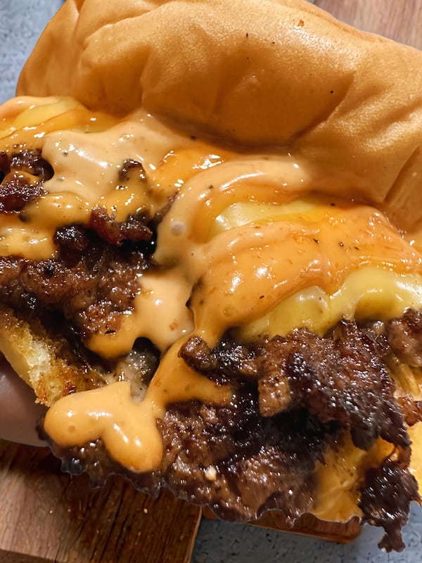 Two burger patties are on a smooth bun. An impossible amount of at least two different kinds of cheeses is oozing out of the bun and off the burger.