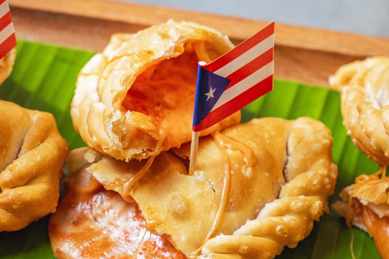 A pizza empanadilla is torn in half and the cheesy filling is oozing out of it. It sits on a banana leaf with a Puerto Rican flag on a toothpick inserted into the crust.