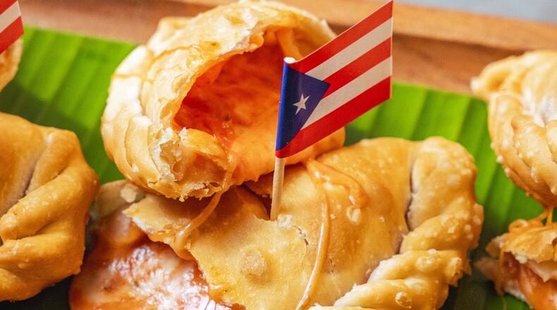A pizza empanadilla is torn in half and the cheesy filling is oozing out of it. It sits on a banana leaf with a Puerto Rican flag on a toothpick inserted into the crust.