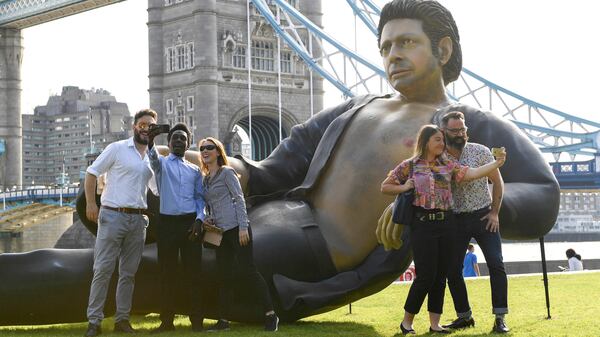 People take photos by a 25ft statue of actor Jeff Goldblum in a pose from a scene in the first Jurassic Park movie, which has been created by a TV channel to celebrate the film's 25th birthday, at Potters Fields Park, London, Wednesday July 18, 2018. Tower Bridge in the background.