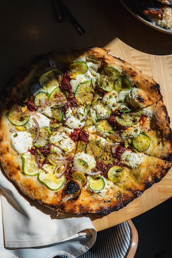 A whole wood-fired, scliced zucchini pizza on a pizza peel, with a napkin tucked under an edge of the pizza.