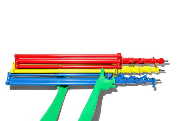 Two hands wearing green gloves hold up three photography light stands in bright primary colors on a white background.