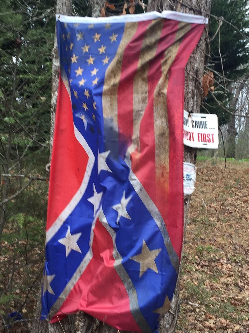 An American flag merged with a Confederate battle emblem hangs from a couple of trees along a highway in North Georgia. It is accompanied by a sign that says, “Fight crime … Shoot first.”