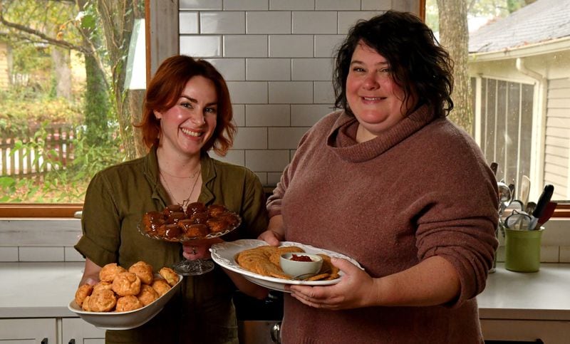 Ashley Thomas (left) and Morgan Perkins, co-owners of Galette, pose with a few of their holiday party bites: Gougeres (left), Sticky Toffee Pudding Bites (center) and Cheddar Black Pepper Sables (right). They are in Thomas' Atlanta home kitchen, where they create the baked goods they sell at local farmers markets. (Styling by Ashley Thomas and Morgan Perkins / Chris Hunt for the AJC)
