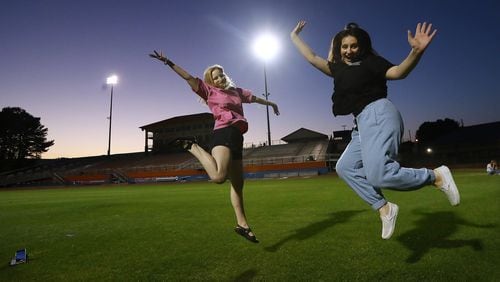 Graduating seniors Megan Hollister (left) and Caitlyn Bradley jump while setting their cell phones on the grass and filming a video of themselves in the Big Orange Jungle and Hugh Buchanan Field as it is lit up to symbolize the class of 2020 is a light to the community at Parkview High School on Tuesday, April 14, 2020, in Lilburn. Every week night at 8:20 pm (20:20 military time) Athletic Director Nick East turns on the scoreboard and stadium lights for 20 minutes and 20 seconds to honor the class of 2020.